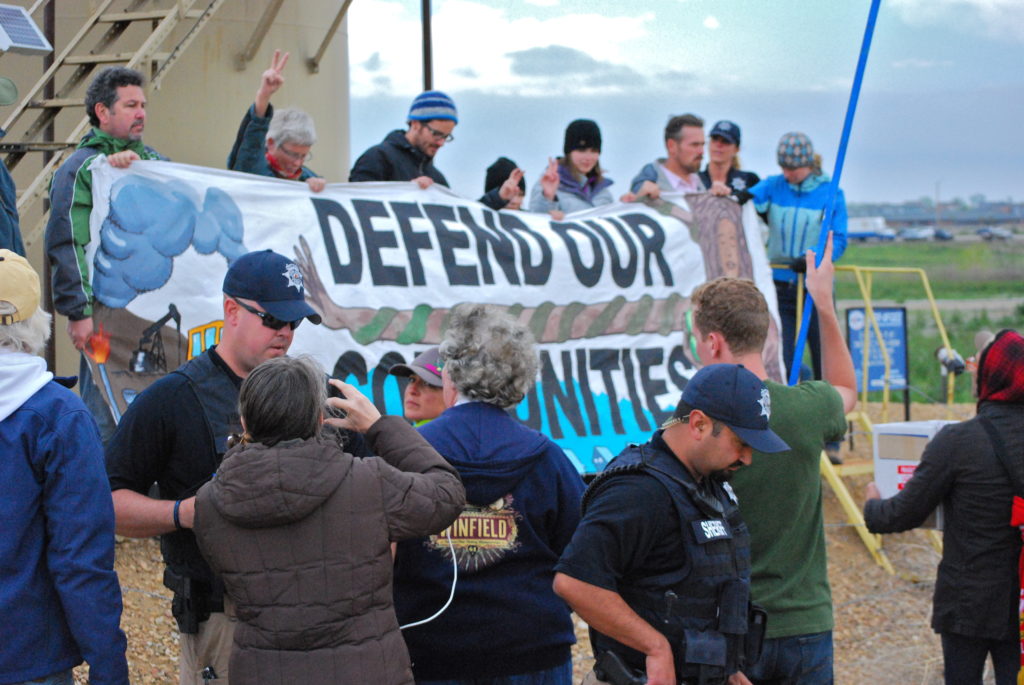 After once again ignoring police demands, protesters unfurled their banner on top of the dirt barriers. As police backup arrived in seven unmarked cars, officers once again drew a new line in the sand, telling protesters if they crossed the barbed wire fence into the actual tank area, they would be arrested. At that point, several protesters crossed the fence while shouting and daring to police to arrest them and accusing police of being afraid to arrest them.