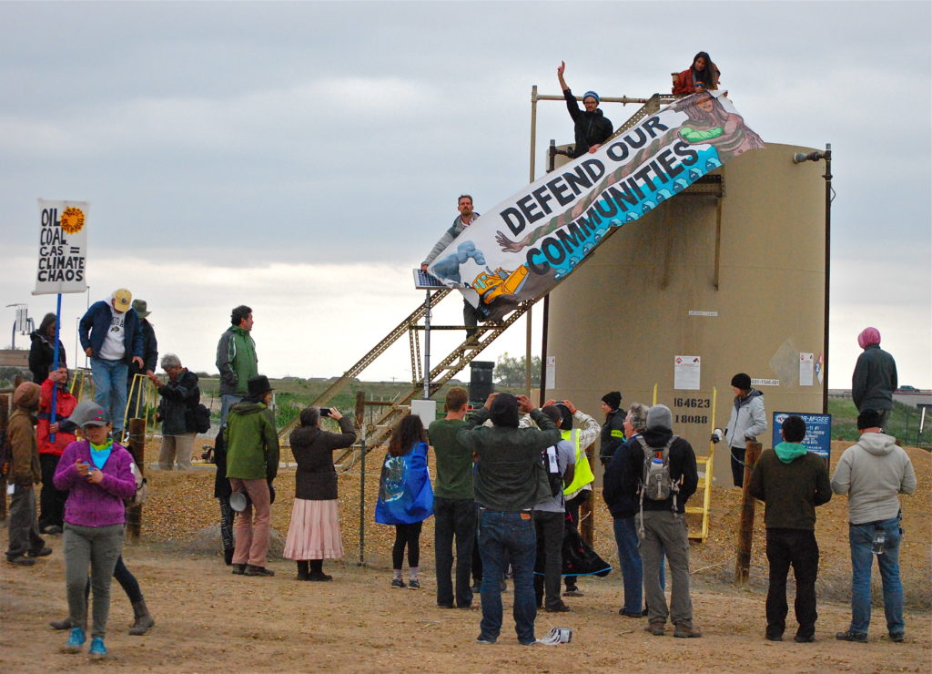 At that point, all of the police got into their cars and drove away together regrouping back on the main road where the majority of protesters were still positioned. As the police were leaving, protesters climbed to the top of the tank amid cheers and attached their banner. 